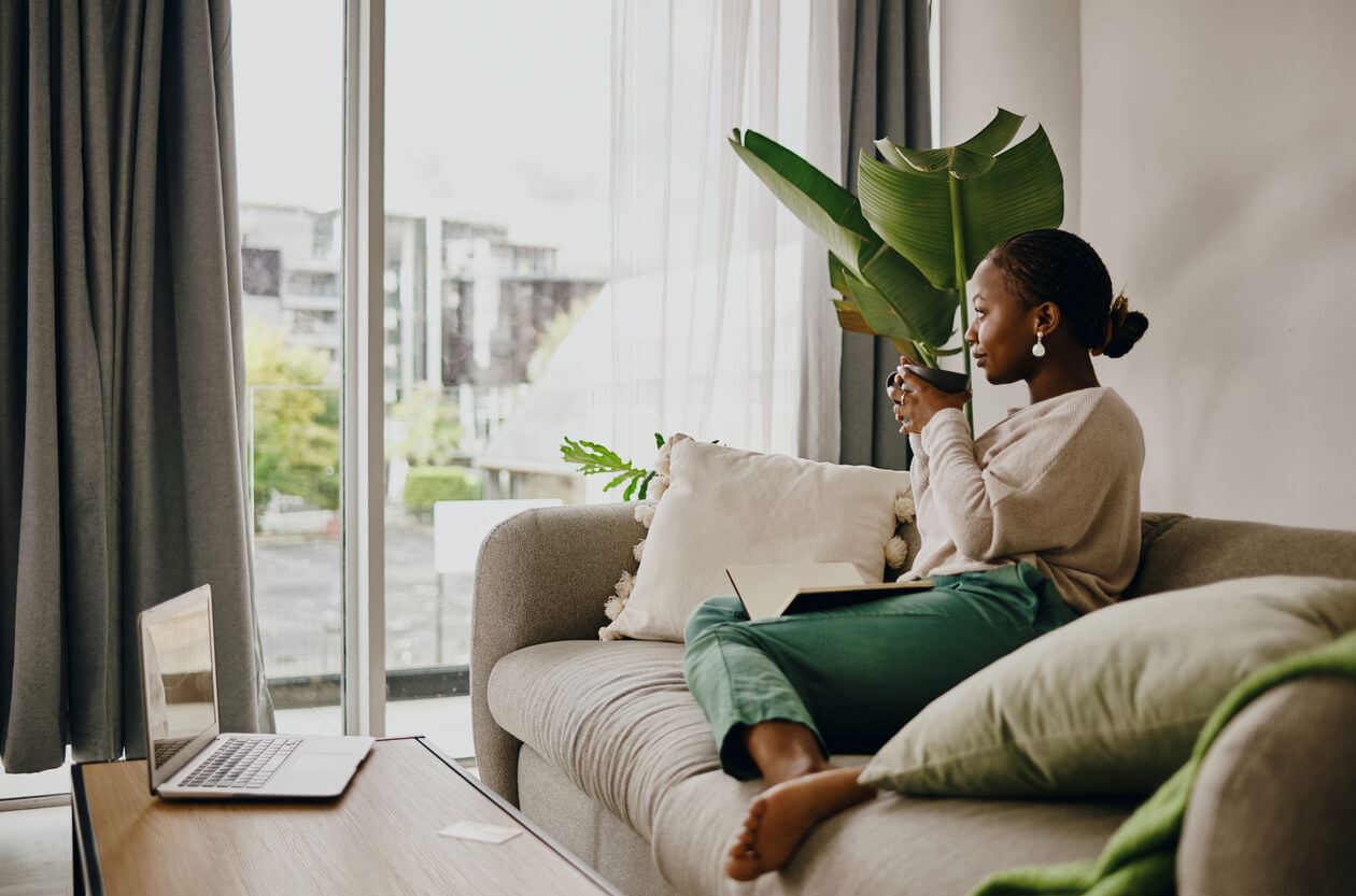 Woman sitting on couch with mug in her hand while she looks outside window.