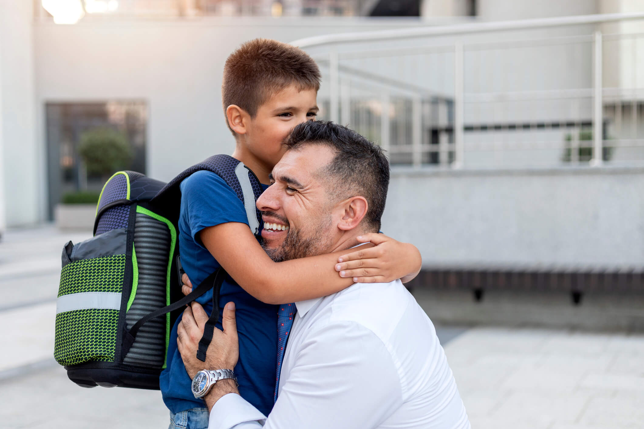 Father hugging son with backpack on in front of school