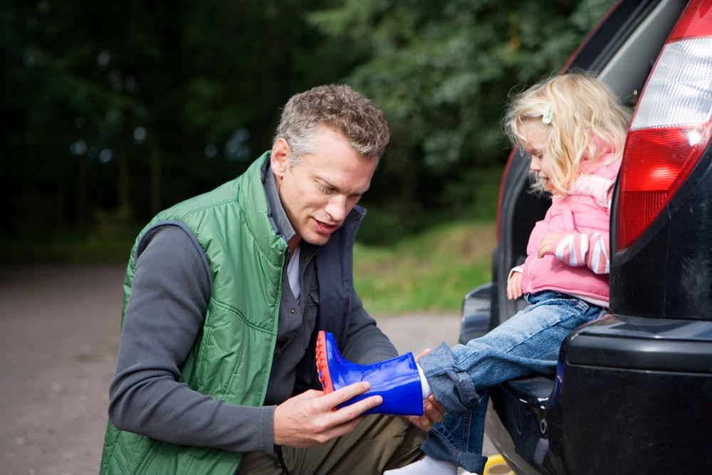 Father putting wellington boot on child foot for countryside walk as she sits in car trunk